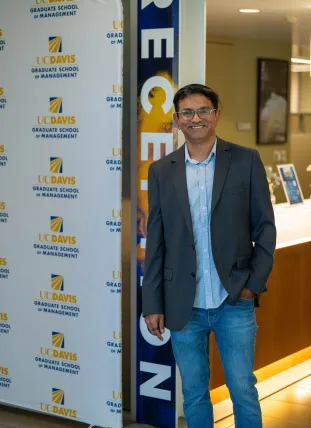 A man in a blazer and jeans stands smiling beside a UC Davis Graduate School of Management banner in an indoor setting.
