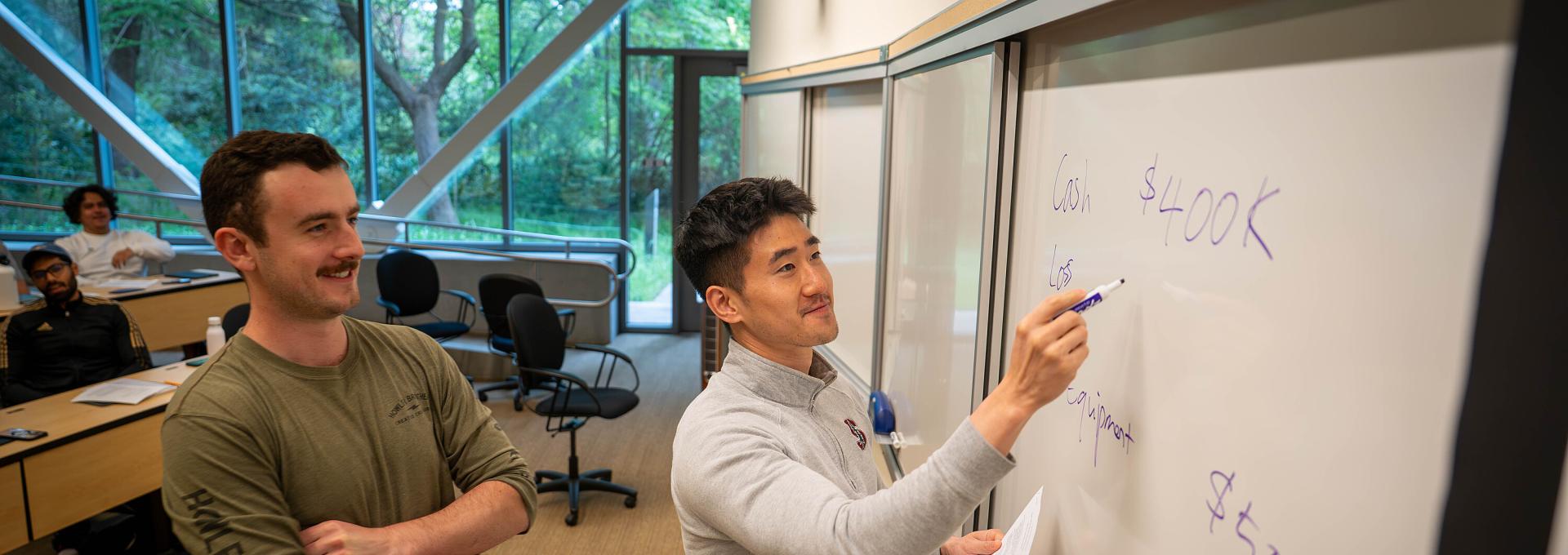 Two students at the front of a GSM classroom writing on a whiteboard