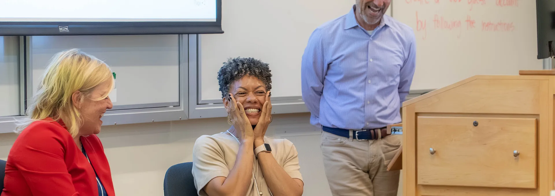 Three people sit and stand at the front of a classroom; the woman in the center smiles with hands on her cheeks, while the others look in her direction and smile.