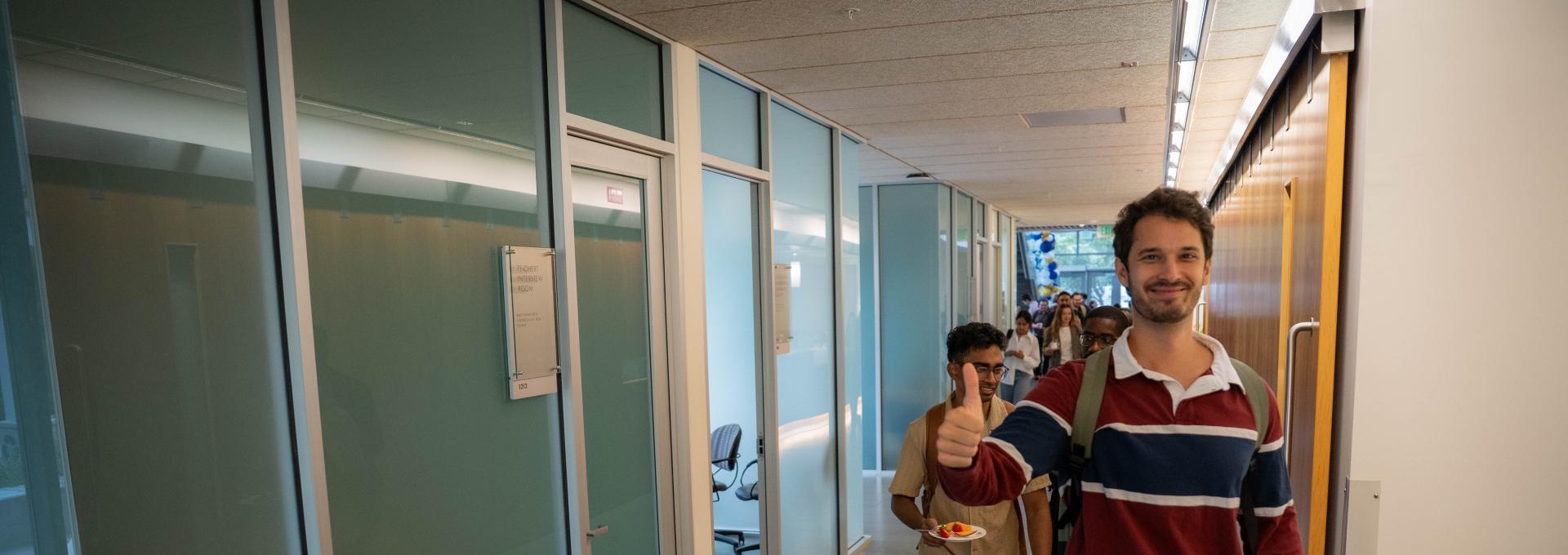 A man in a striped sweater walks down a hallway giving a thumbs up, followed by another person; offices with glass walls line the corridor.