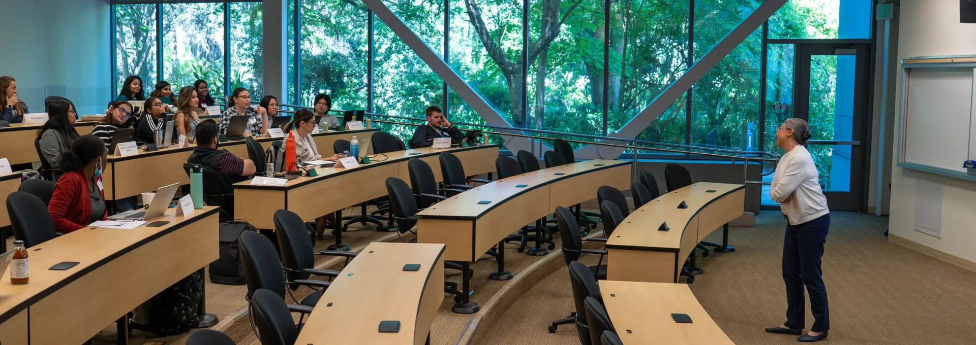 A person stands at the front of a modern classroom lecturing to students seated at curved desks, with large windows showing greenery outside.