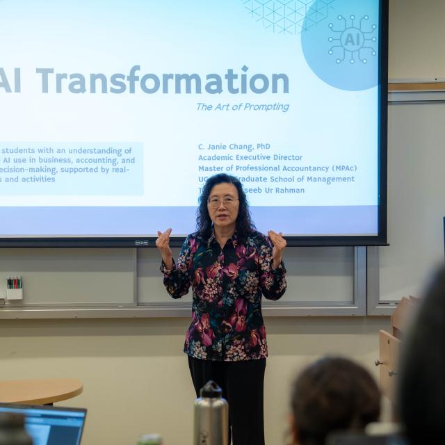 Professor Janie Chang stands in front of a projected presentation titled "AI Transformation" in a classroom, speaking to an audience.