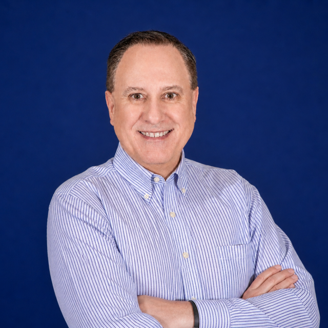 Tim Akin in a light blue striped shirt stands with arms crossed and smiles at the camera, set against a plain dark blue background.