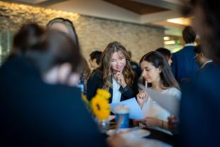 Two MBA students in professional attire review a document together at a networking event, surrounded by other attendees. A sunflower centerpiece and notepads are visible on the table.