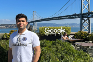 Talha Amin stands on an outdoor terrace at Google’s San Francisco office, with the Bay Bridge and blue sky in the background. He’s wearing a white polo shirt, light pants and sunglasses, surrounded by lush green plants.
