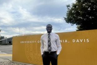 A man in formal attire stands in front of a yellow University of California, Davis sign under a partly cloudy sky.