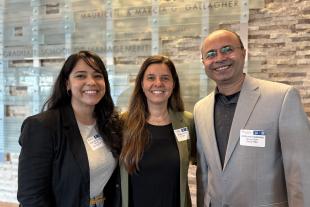 Three people stand indoors, smiling at the camera. They wear business attire and conference name tags. A wall sign behind them reads "Graduate School of Management.