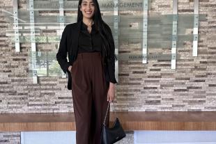 A woman stands indoors in business attire, holding a handbag and smiling, in front of a wall displaying names and a sign reading “Graduate School of Management.”