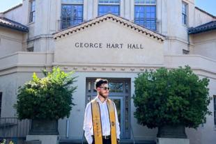 A man wearing a graduation stole stands in front of George Hart Hall on a sunny day.