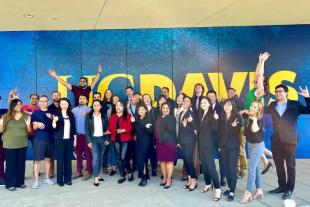 A group of people posing and smiling in front of a large blue and yellow “UC DAVIS” sign, with several making enthusiastic gestures.
