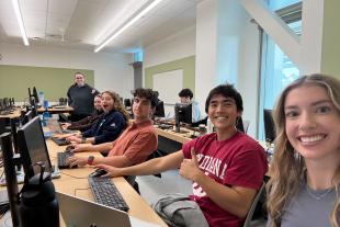 A group of students sit at desks with computers in a classroom, smiling at the camera; an instructor stands at the back of the room.