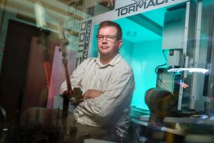 A man wearing glasses and a striped shirt sits with arms crossed in front of a Tormach CNC machine in a workshop.