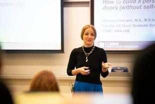 A woman stands in front of a classroom giving a presentation, gesturing with one hand. A projection screen and audience members are visible.