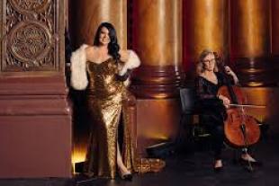 A woman in a gold sequin gown stands next to a column, while another woman sits nearby playing a cello.