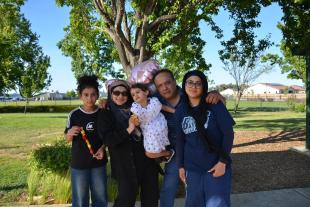A family of five poses together outdoors in a park on a sunny day, with trees, grass, and houses in the background. One member holds heart-shaped balloons.