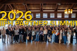 A large group of people standing together indoors, smiling and waving, with “2026 IIFH Partner Summit” in large yellow text above them.
