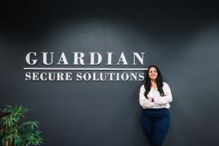 A woman stands with arms crossed in front of a dark wall displaying the words "Guardian Secure Solutions.