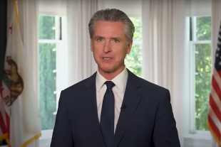 Gavin Newsom in a navy suit and tie stands indoors between two windows, with American and California state flags visible in the background.
