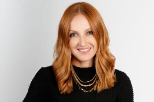 Woman with long red hair wearing a black textured top and layered gold necklaces, seated and smiling against a plain light background.