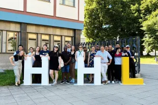 A group of people pose outdoors, smiling and standing behind large white and yellow letters spelling "HHL" on a paved area near a building and trees.