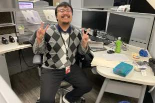 Man sitting at an office desk, smiling and making peace signs with both hands. Multiple monitors, papers, and office supplies are visible around him.