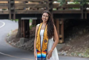 Woman in a white dress and graduation stole stands on a road in front of a wooden bridge, smiling at the camera.