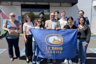 A group of people stand outdoors holding a blue UC Davis Aggies flag; one person holds a box, and an American flag is visible in the background.