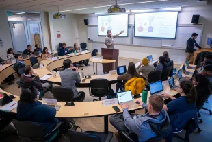 A lecturer presents to students seated in a semicircular classroom, some taking notes on laptops, with slides projected on screens at the front.