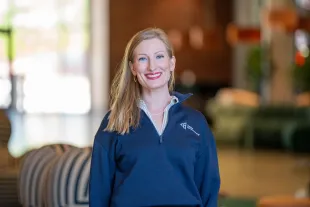 Jaimie Asakawa smiling, wearing a navy pullover and pearl necklace, standing indoors with blurred background.