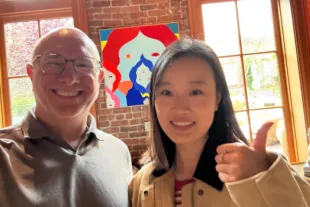 Two people smile at the camera indoors; one gives a thumbs up. A colorful abstract painting and large windows with a garden view are in the background.