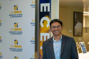 A man in a blazer and jeans stands smiling beside a UC Davis Graduate School of Management banner in an indoor setting.