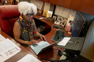 Woman with white curly hair works at a desk with a laptop and monitor, surrounded by paperwork and framed photos.