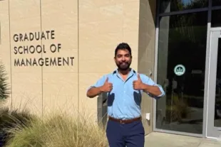 A man in a light blue shirt and dark trousers stands in front of the Maurice J. Gallagher Graduate School of Management building giving two thumbs up.