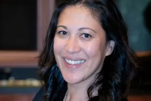 A woman with dark hair smiling at the camera, wearing a black shirt and a gold necklace.