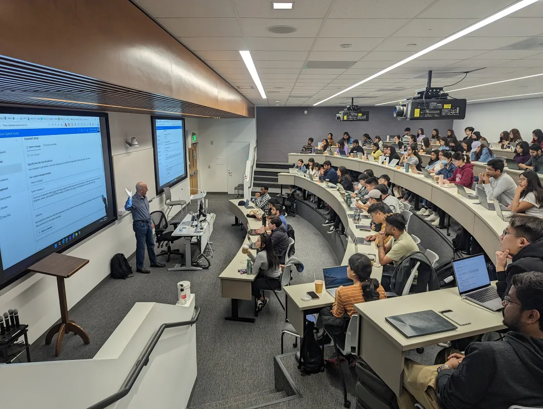 A professor stands at the front of a tiered lecture hall, presenting slides to students seated at desks with laptops and notebooks.