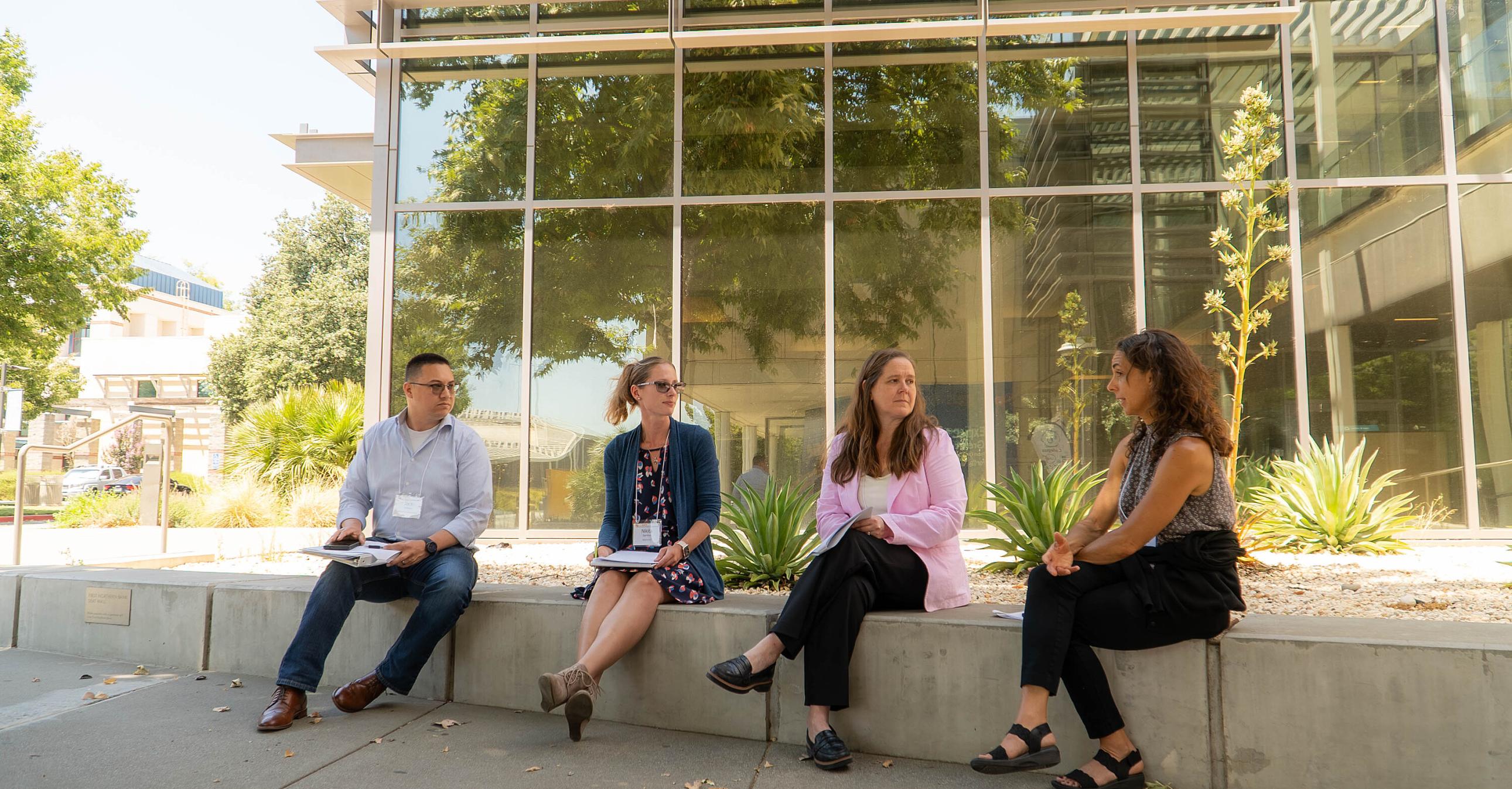 Four people sit on a low concrete wall outside Gallagher Hall, having a discussion with notebooks in hand, on a sunny day.