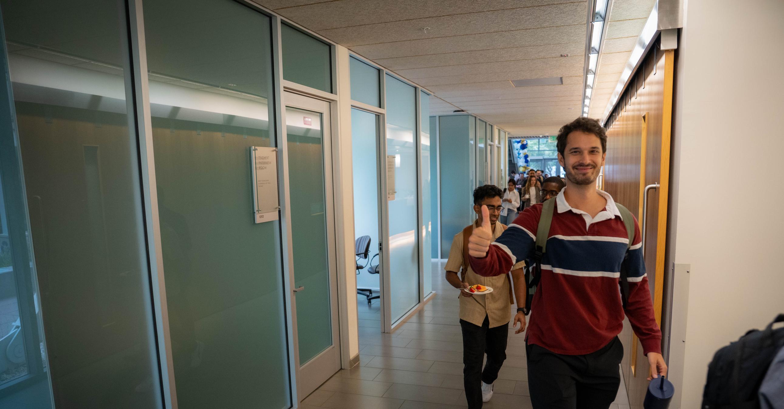 A man in a striped sweater walks down a hallway giving a thumbs up, followed by another person; offices with glass walls line the corridor.