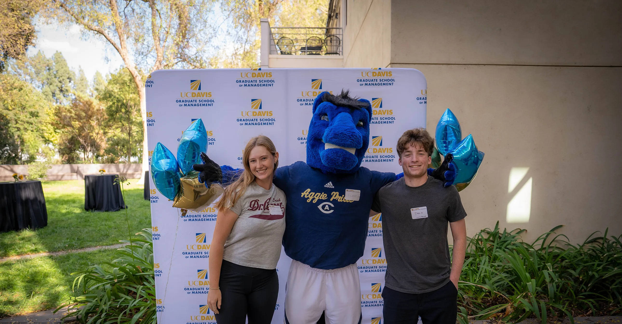 Two people stand beside a blue horse mascot holding star-shaped balloons, posing in front of a UC Davis Grandparents Day banner outdoors.