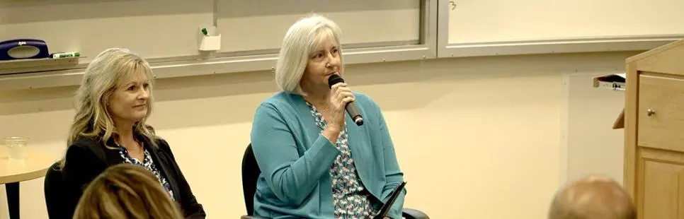 Two women sit at the front of a classroom; one speaks into a microphone while the other listens. Several people are seated facing them.