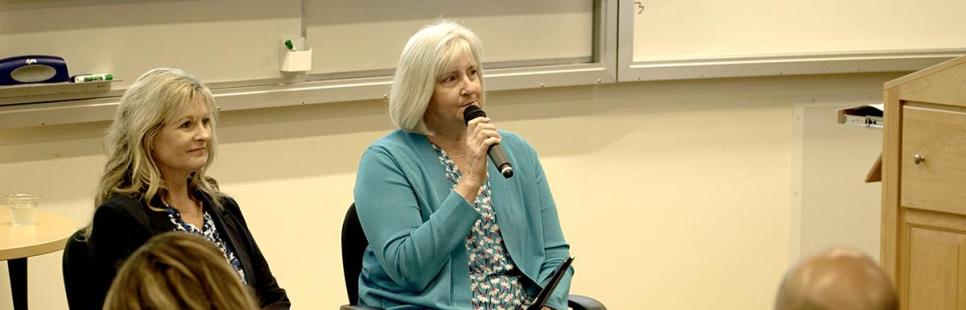 Two women sit at the front of a classroom; one speaks into a microphone while the other listens. Several people are seated facing them.