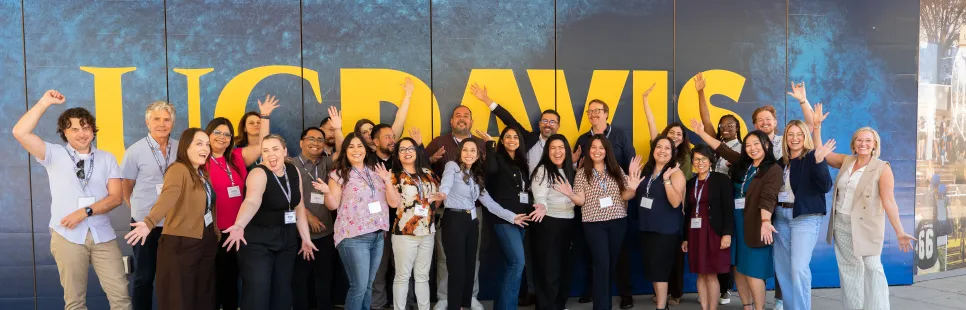 A group of people pose and smile in front of a blue and gold UC Davis sign outside a building on a sunny day.