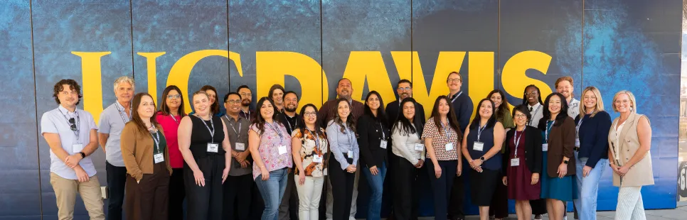 A group of people stands in front of a large UC Davis sign on a blue and yellow background, posing for a group photo outdoors.