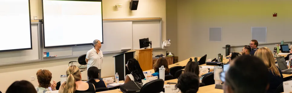 A person stands at the front of a classroom giving a presentation to an audience seated at desks, facing a screen with slides projected.