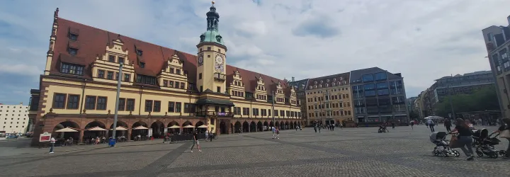 Large cobblestone square with a historic building featuring a clock tower on the left, modern buildings on the right, and people walking and sitting around.