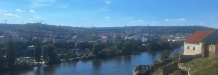 A river runs through a city with buildings and cranes, bordered by greenery and a hillside with a small structure under a blue sky.