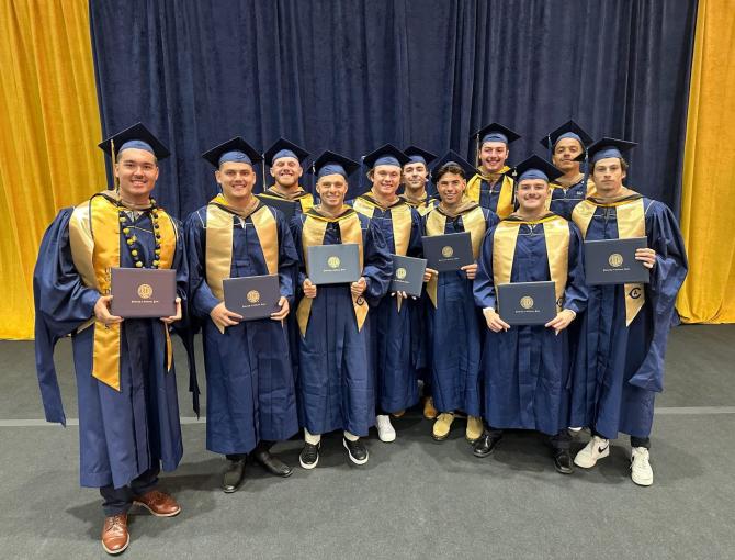 Twelve graduates in blue and gold caps and gowns stand together, holding diplomas and smiling in front of gold and navy curtains.