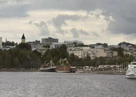 Harborfront in Lappeenranta, Finland, with boats docked along the shoreline and apartment buildings rising behind under a cloudy summer sky.