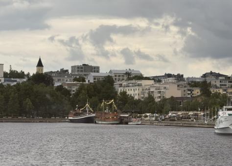Harborfront in Lappeenranta, Finland, with boats docked along the shoreline and apartment buildings rising behind under a cloudy summer sky.