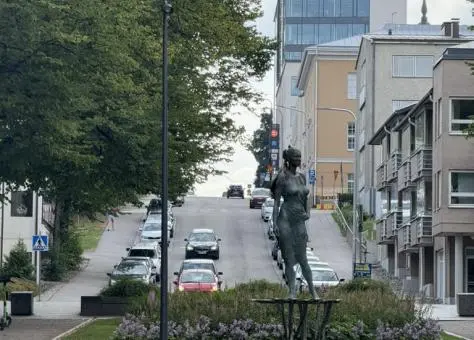 Street view in Lappeenranta, Finland, featuring a bronze statue in the foreground, flowerbeds, and a tree-lined road leading uphill past modern and historic buildings.
