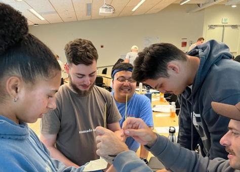 Five people work together to build a tall structure using uncooked spaghetti and marshmallows in a classroom setting.